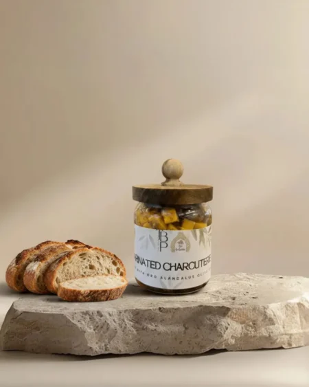 Glass jar of marinated charcuterie made with Oro Al Andalus olive oil, served on a travertine stone slab alongside sliced sourdough bread