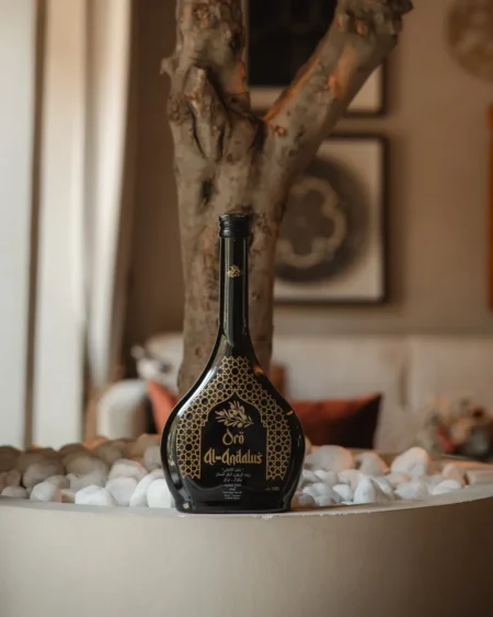 Oro Al Andalus Arabesca Extra Virgin Olive Oil 500ml black bottle with gold Andalusian arch design, displayed on white pebbles beside a natural olive tree trunk in a luxury interior setting