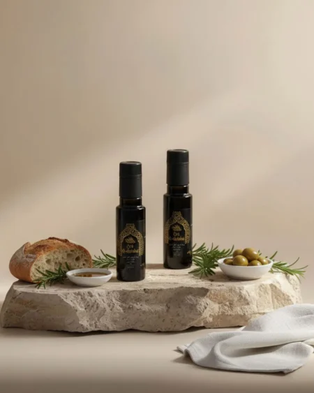 Two black Oro Al Andalus Essence EVOO 100ml bottles on a travertine stone slab with fresh rosemary, green olives, a dipping bowl and artisan sourdough bread