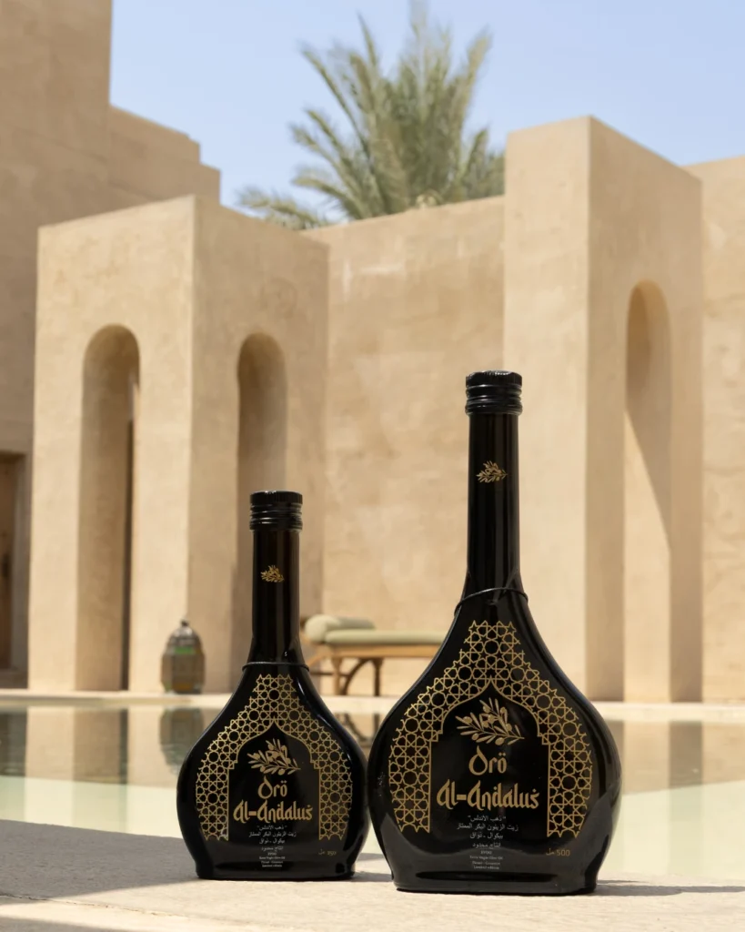 Two Oro Al Andalus Arabesca Extra Virgin Olive Oil bottles in 250ml and 500ml displayed on a stone ledge at a luxury desert resort with Moorish arches and a palm tree in the background