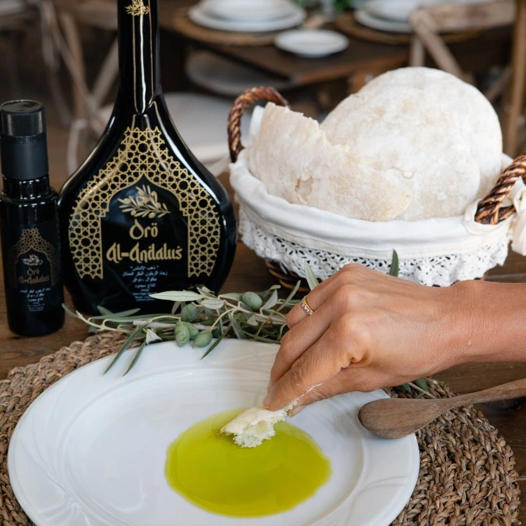 Hand dipping artisan bread into golden Oro Al Andalus Extra Virgin Olive Oil on a white plate, with Arabesca 500ml and Essence 100ml bottles, fresh olive branch and bread basket on a rustic dining table