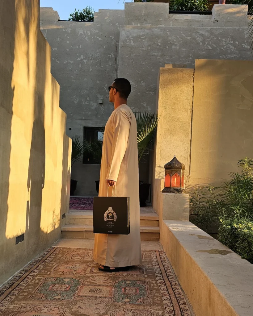 Man in white kandura holding an Oro Al Andalus luxury gift box in a traditional Arabic home courtyard with a glowing Moorish lantern at golden hour